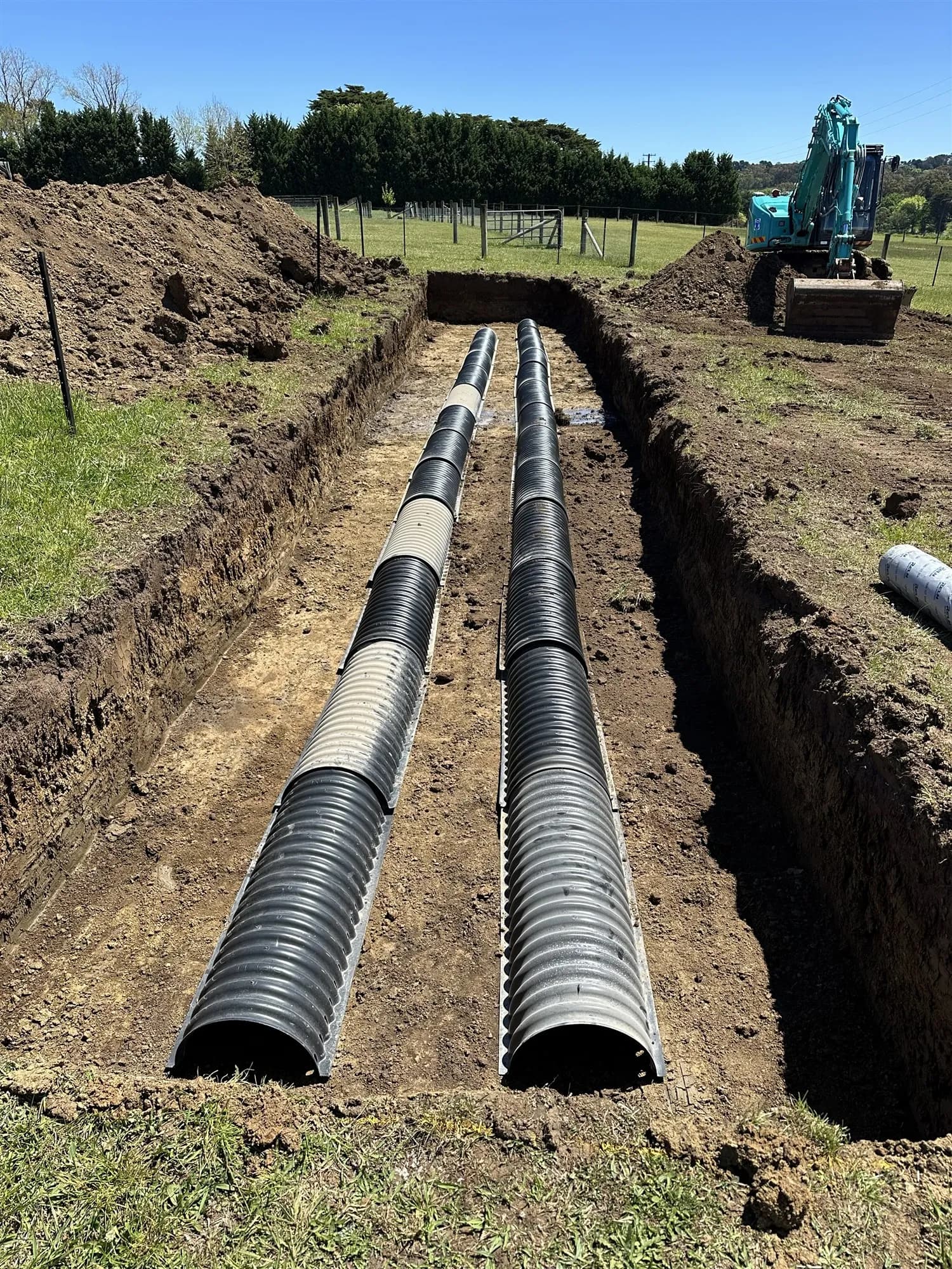 Twin corrugated stormwater pipes laid in a long trench, with an excavator behind