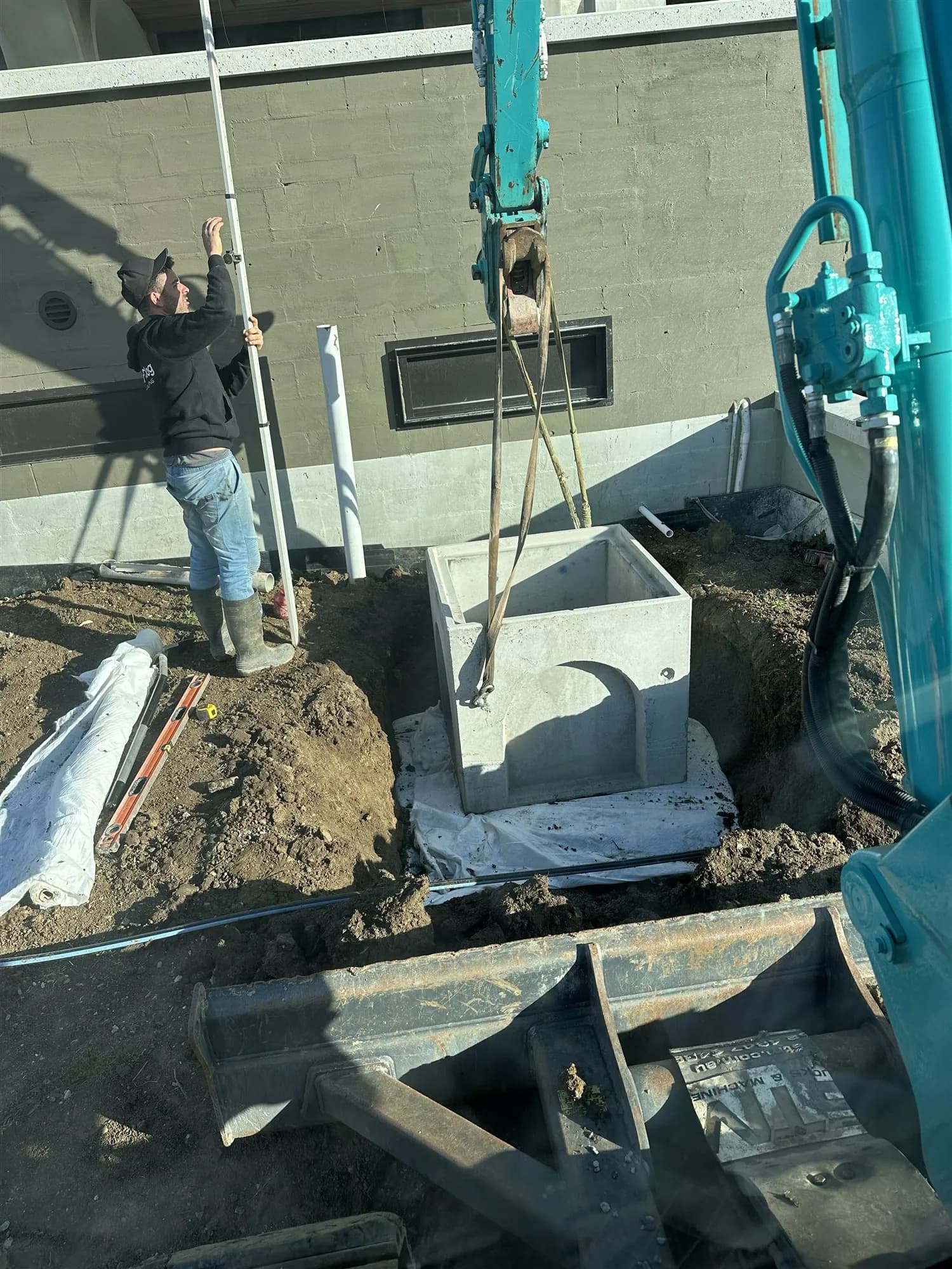 Concrete stormwater pit being lowered into place by an excavator beside a new house