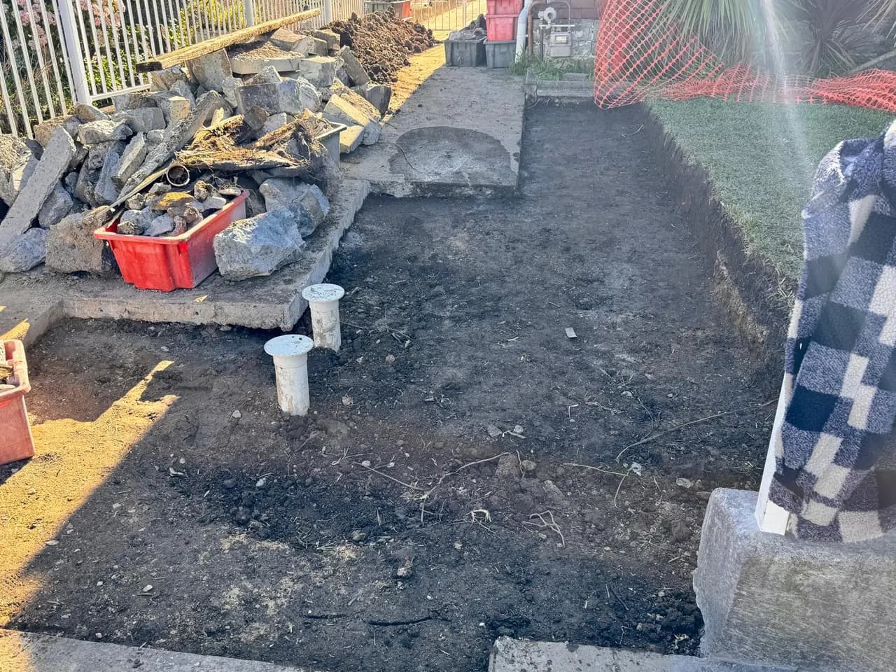 Open drainage excavation across a Sydney yard with new inspection risers standing proud, broken concrete stacked to one side and orange safety mesh around the dig