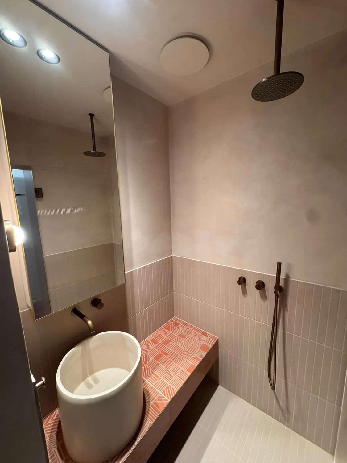 Completed ensuite in pink microcement and fluted tile, with aged brass tapware, a ceiling-mounted rain head and a vessel basin on a tiled bench