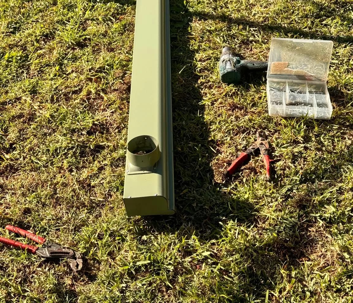 New Colorbond gutter length laid out on the lawn with the downpipe outlet fitted, alongside tin snips, screws and a drill