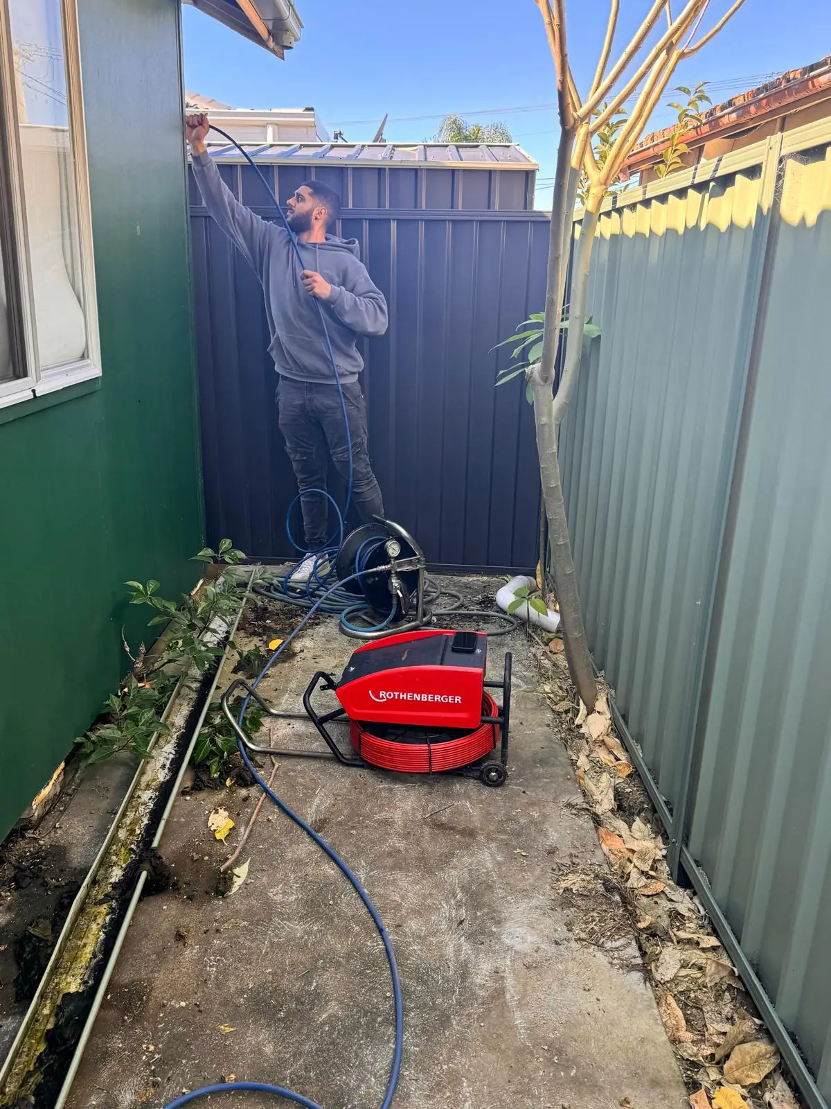 Mr. Clog plumber feeding a hose up a narrow side passage beside a house, with a drain machine and an open trench along the wall
