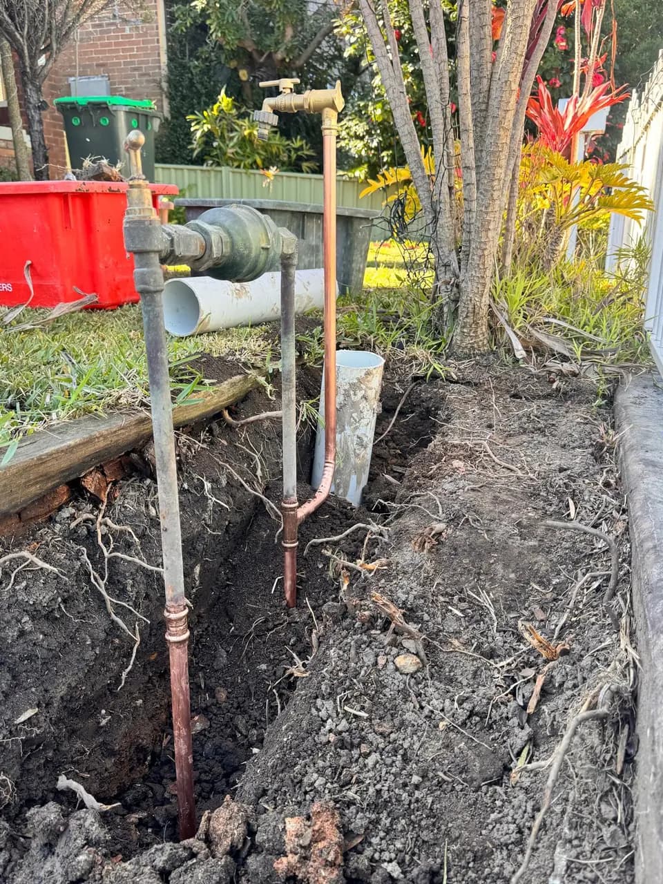 Old galvanised water service pipe beside its new copper replacement in an open trench, with a new brass garden tap fitted