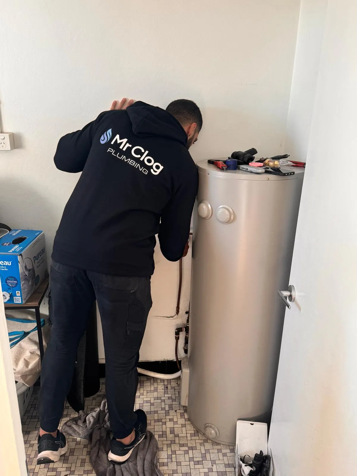 Mr. Clog plumber in a branded hoodie connecting a new stainless hot water cylinder beside the old unit it replaces